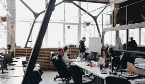 A team sitting at the tables with computers and laptops and listening to the colleague standing by the flipchart in a spacious light modern office