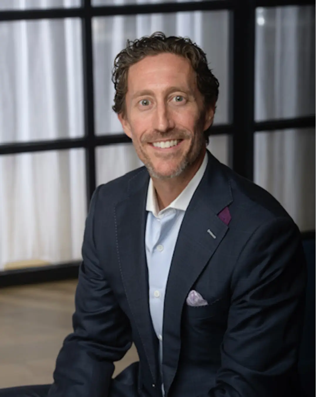 Smiling man in a dark blazer with a white shirt sitting indoors.