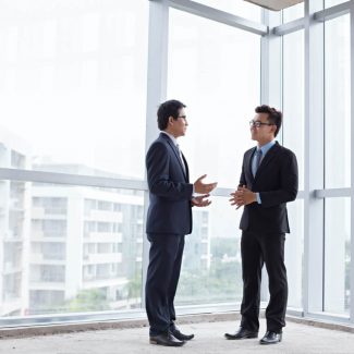 Two men discuss a business sale near the glass wall window of an office corner