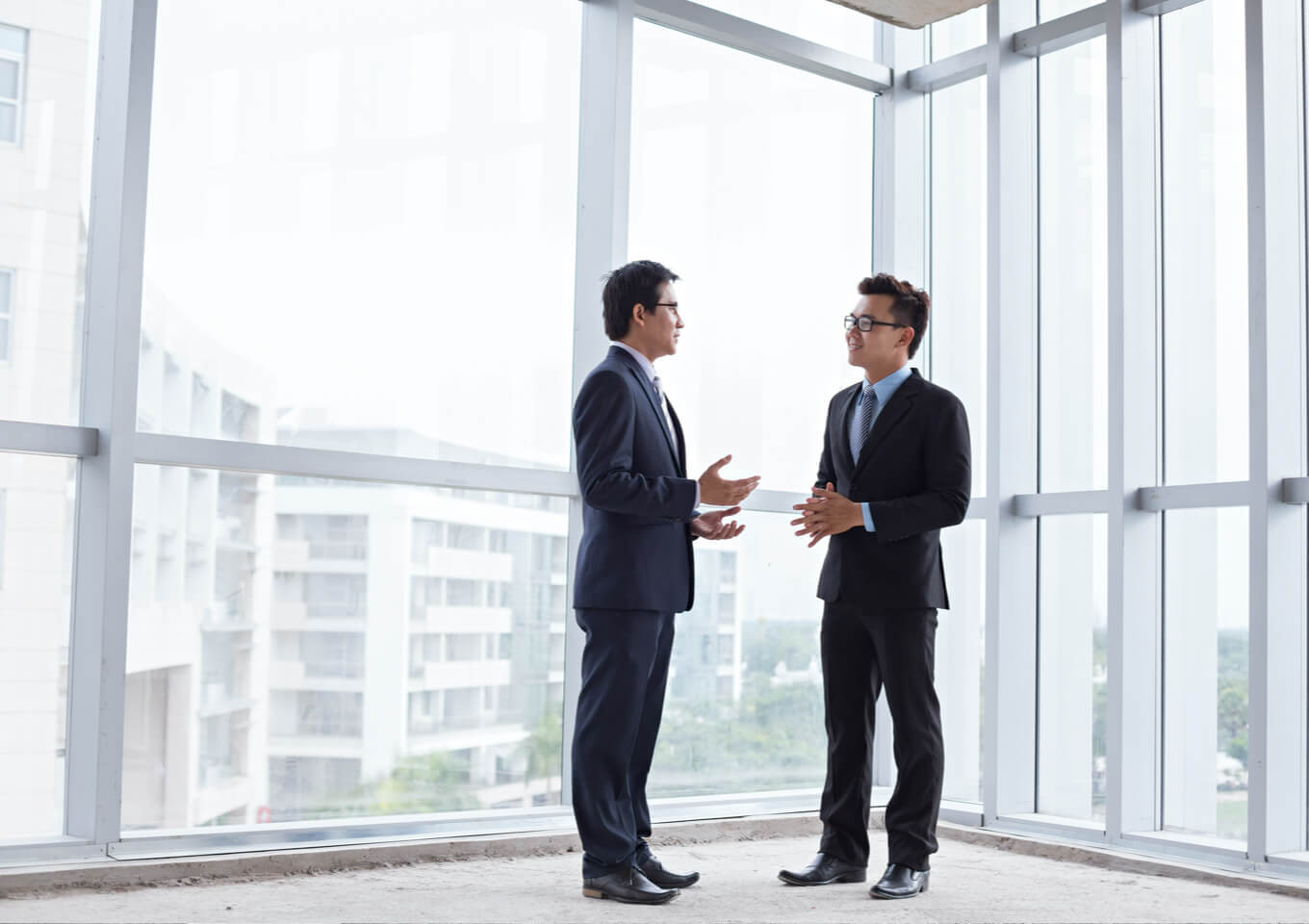 Two men discuss a business sale near the glass wall window of an office corner