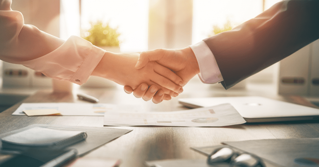 Two people shaking hands over business documents in a bright office.