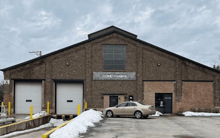 A beige car parked outside a brick industrial building on a cloudy day.