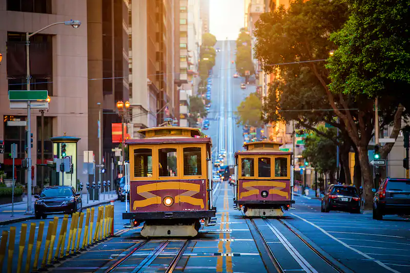Two vintage cable cars ascend a steep, sunlit city street lined with trees and buildings. Two vintage cable cars ascend a steep, sunlit city street lined with trees and buildings.