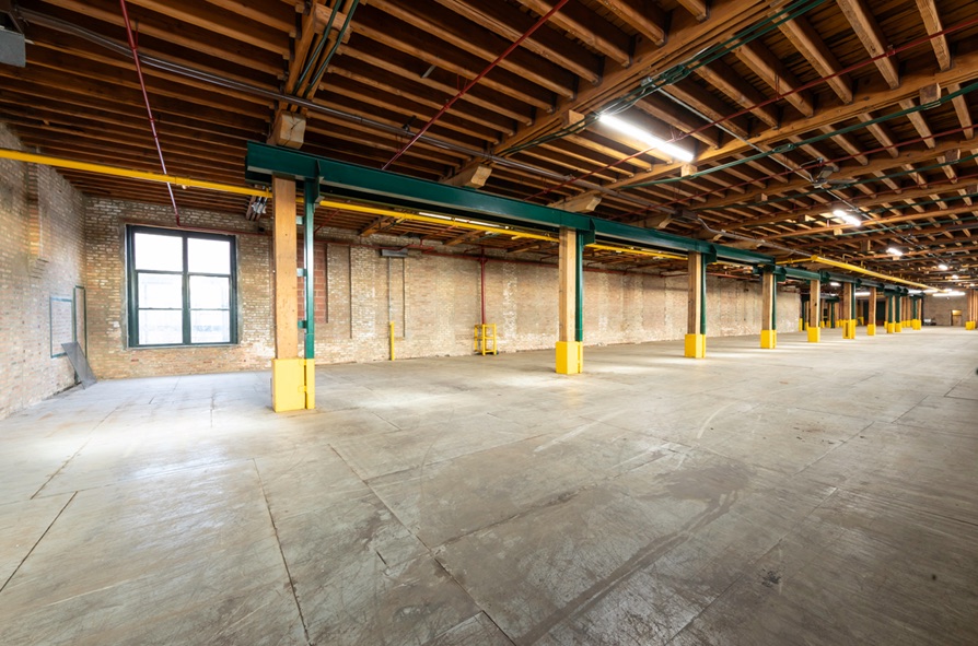 Empty warehouse floor at 2443 W 16th St under exposed timber joists, with painted steel beams and original brick walls